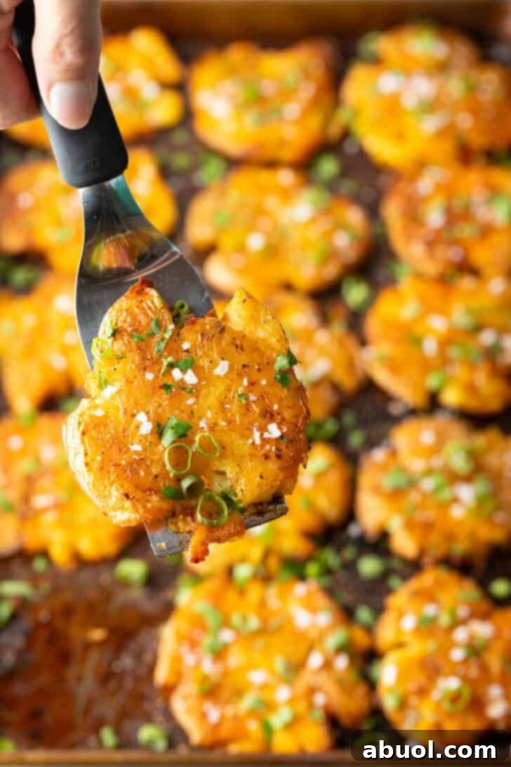 Top down view of 15 golden crispy smashed potatoes arranged on a baking sheet. A hand holding a spatula proudly presents a perfectly cooked smashed potato directly to the camera, highlighting its enticing golden-brown and crispy exterior.