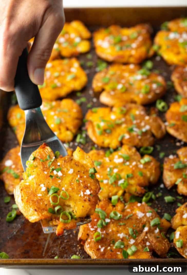 Top down view of 15 golden crispy smashed potatoes perfectly arranged on a baking sheet. A hand with a spatula holds up one of the cooked potatoes, showcasing its crunchy texture and golden finish directly to the viewer.