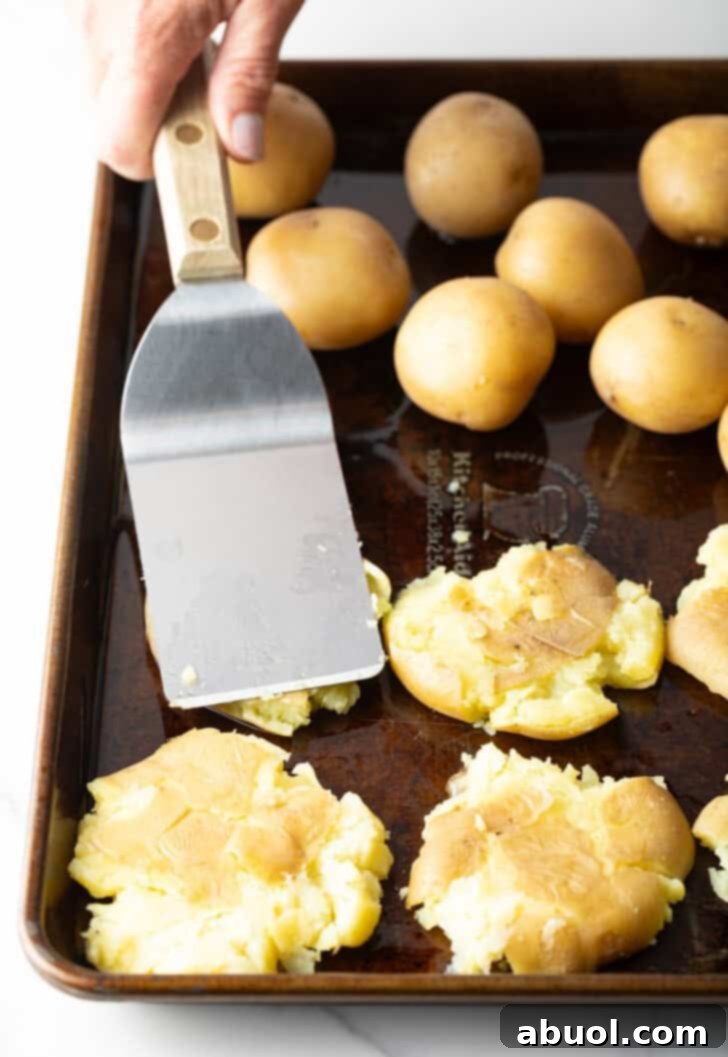 Using a sturdy flat metal spatula to gently but firmly smoosh small, tender boiled yellow potatoes directly onto a baking sheet, forming thin, irregular potato cakes.