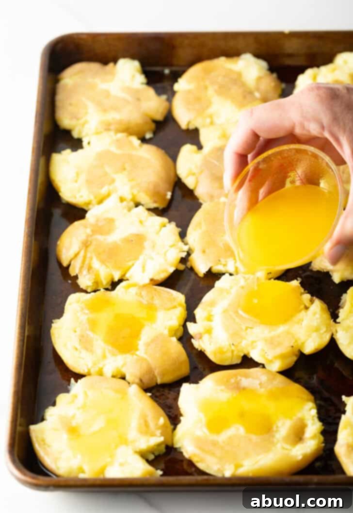 A hand pouring a stream of golden melted ghee from a small glass bowl evenly over the unbaked smashed potatoes arranged on a baking sheet, ensuring each surface is ready for coating.