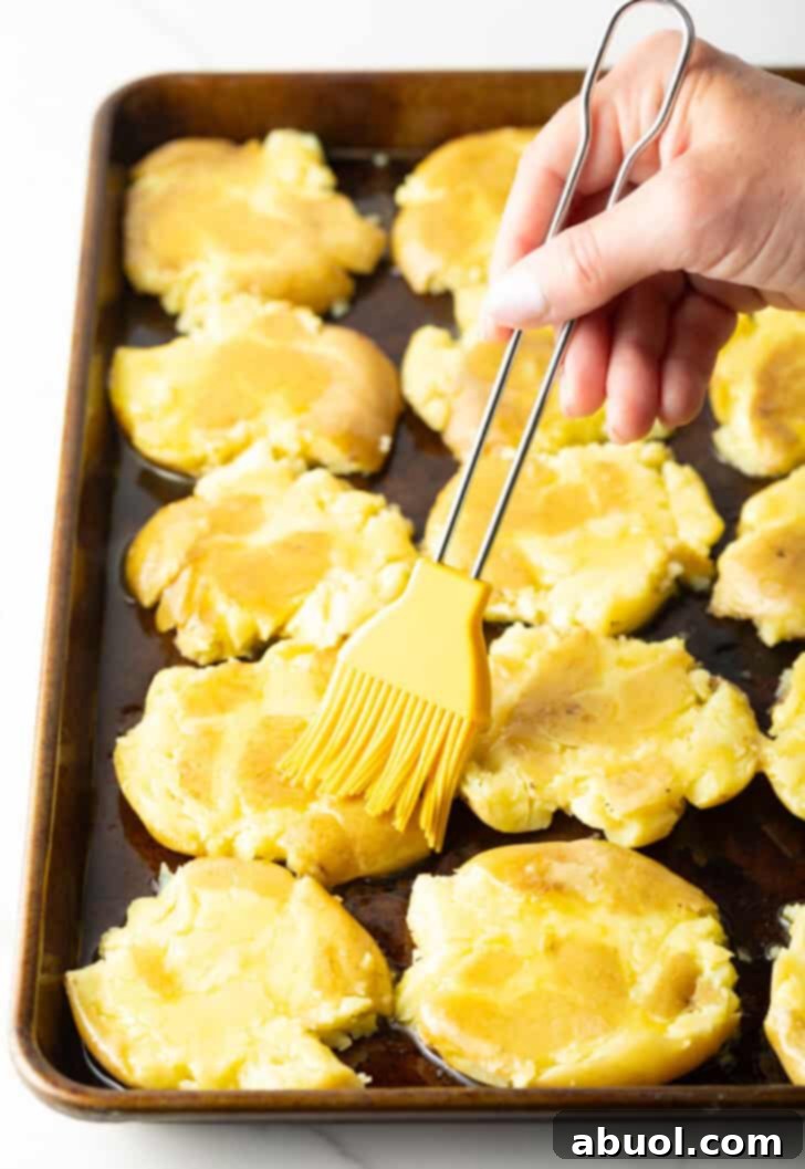 A hand using a yellow basting brush to meticulously spread melted ghee over the tops of the smashed potatoes on a baking sheet, ensuring every nook and cranny is thoroughly coated for maximum crispness.