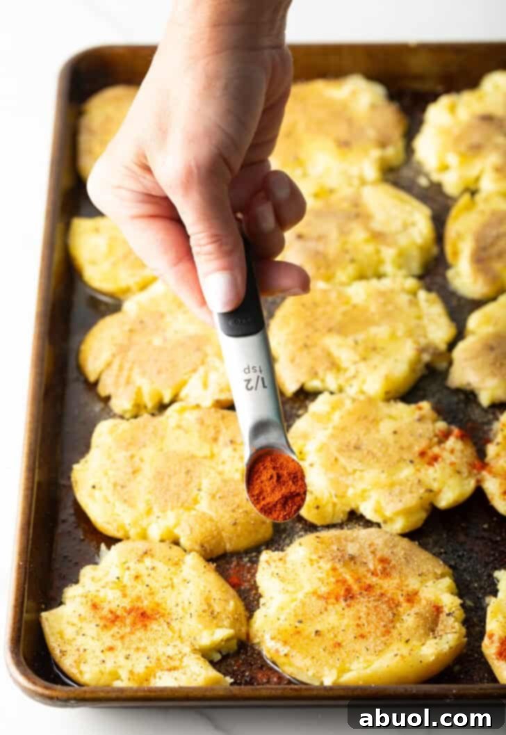 A hand adding a precise amount of spices from a small measuring spoon over the smashed potatoes on a baking sheet, preparing them for their flavorful bake.