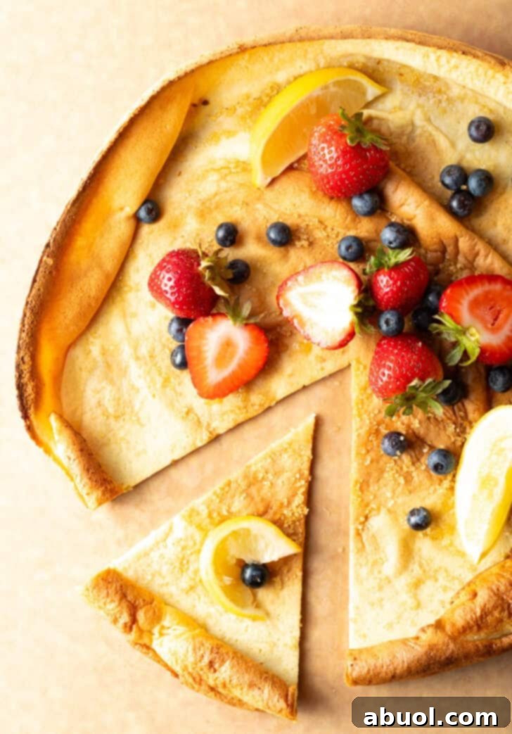Top down view of a Dutch Baby pancake on a cutting board, garnished with fresh strawberries, blueberries, and lemon slices. A slice has been pulled to the side.