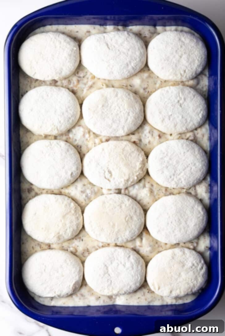 Frozen biscuits lined up neatly on top of a blue pan filled with sausage gravy, ready for baking.