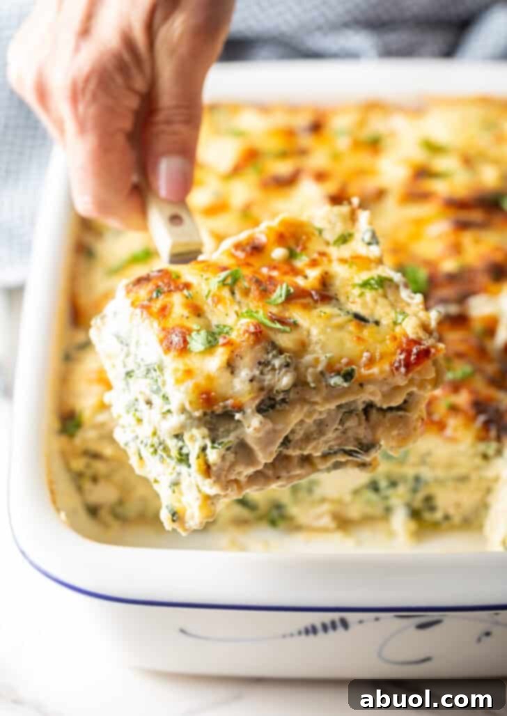 A hand skillfully lifting a generous square of cheesy Chicken Alfredo Lasagna from a white baking dish using a serving spatula.