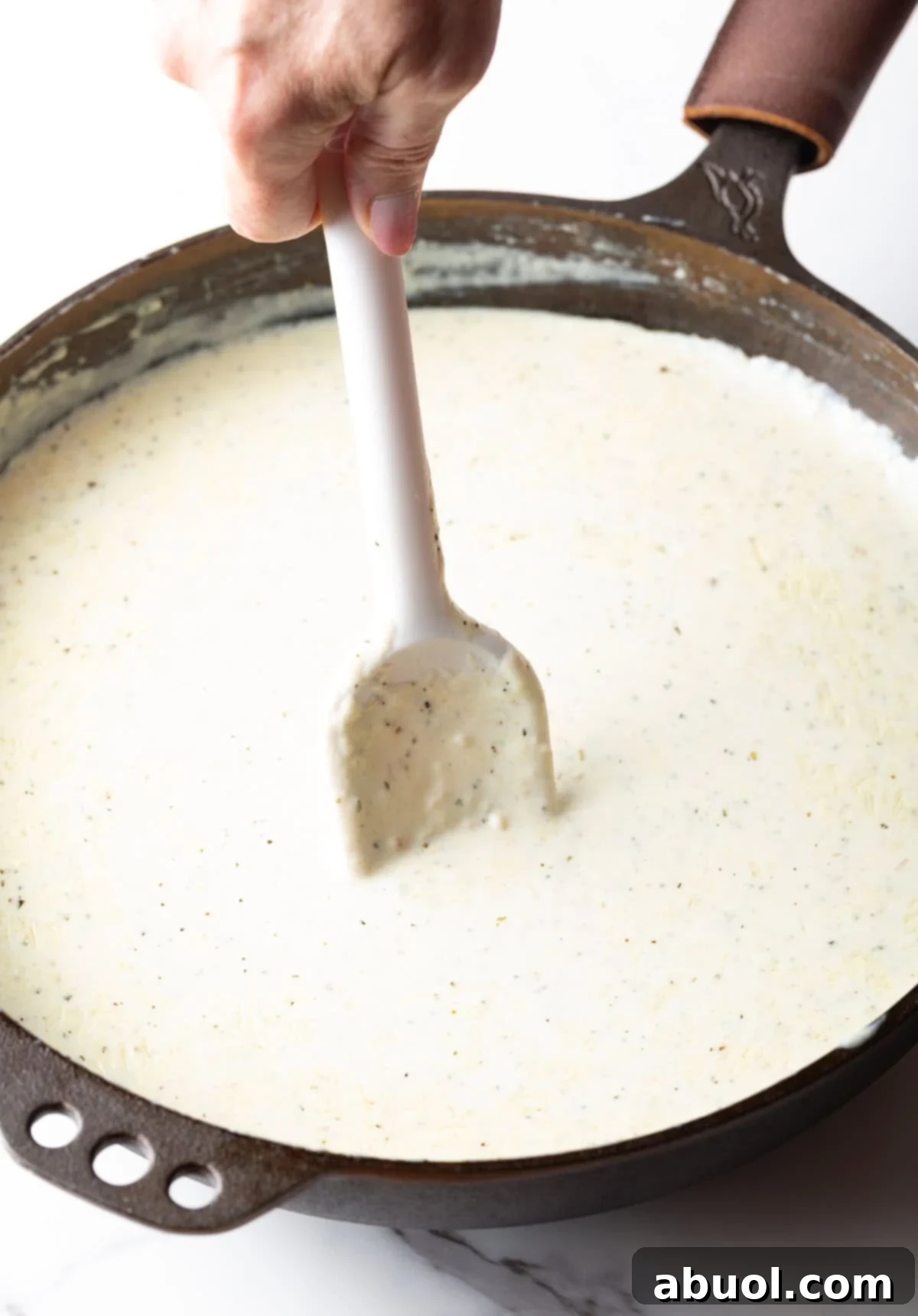 A close-up shot of a hand stirring rich, creamy Alfredo sauce in a large skillet with a white spatula.