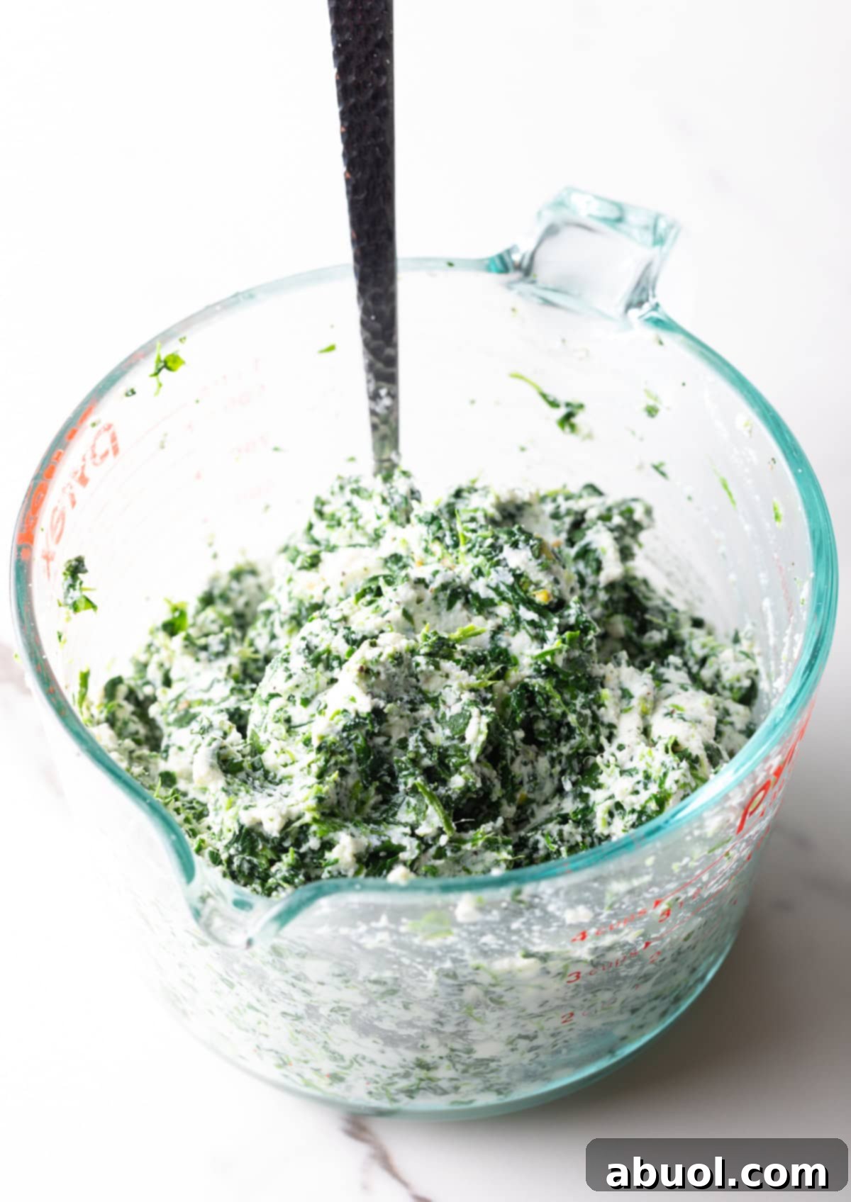 A glass measuring cup holds a mixture of thawed frozen spinach and creamy ricotta cheese, with a metal spoon resting inside.