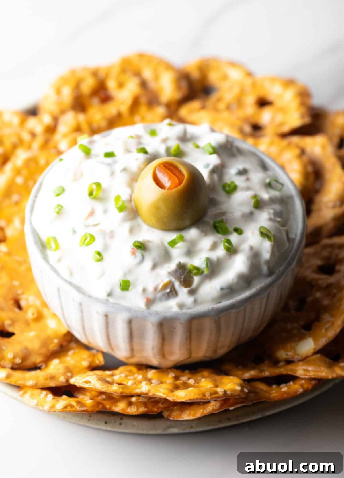 White bowl of dirty martini dip topped with one green olive, surrounded by pretzel chips.