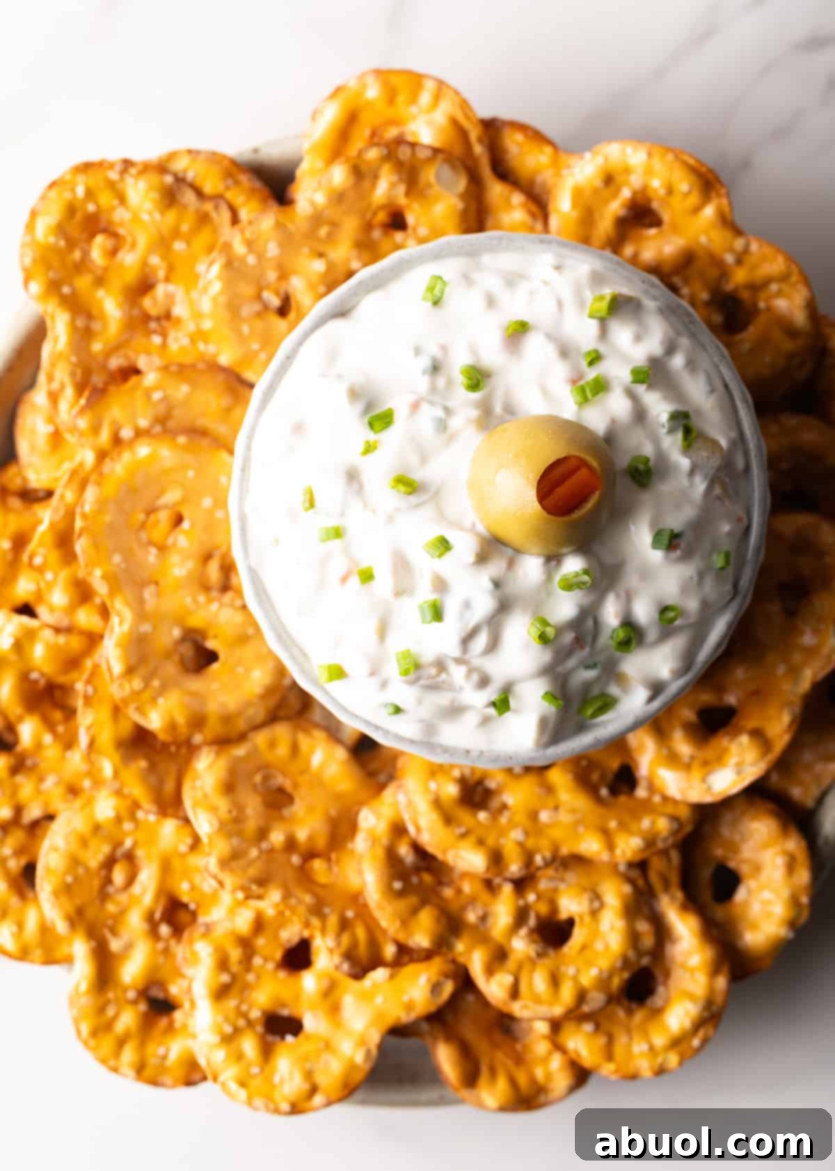 Top down view of a white bowl of dirty martini dip topped with one green olive, surrounded by pretzel chips.