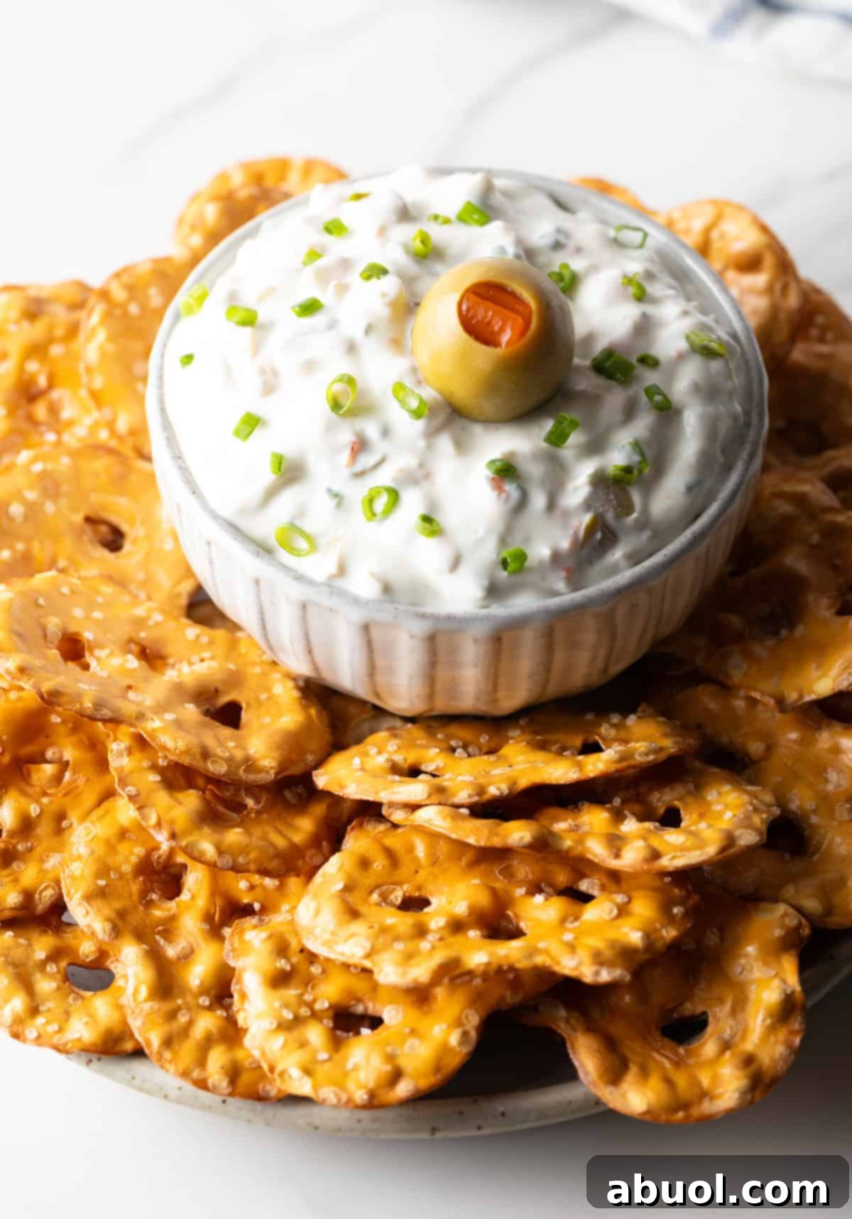 White bowl of dirty martini dip topped with one green olive, surrounded by pretzel chips.