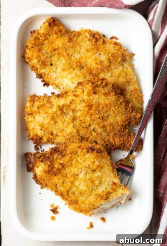 Three perfectly golden-brown baked chicken cutlets resting on a white platter, garnished with fresh herbs, showcasing their crispy texture.