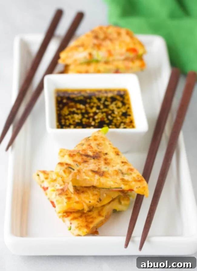 Korean Pancakes #ASpicyPerspective #KoreanPancakes #KoreanPancakeRecipe #Pajun #Pajeon #Korean #Vegetarian Crispy Korean pancakes (Pajeon) cut into wedges and elegantly arranged on a plate next to a small bowl of spicy soy dipping sauce, ready to be enjoyed.