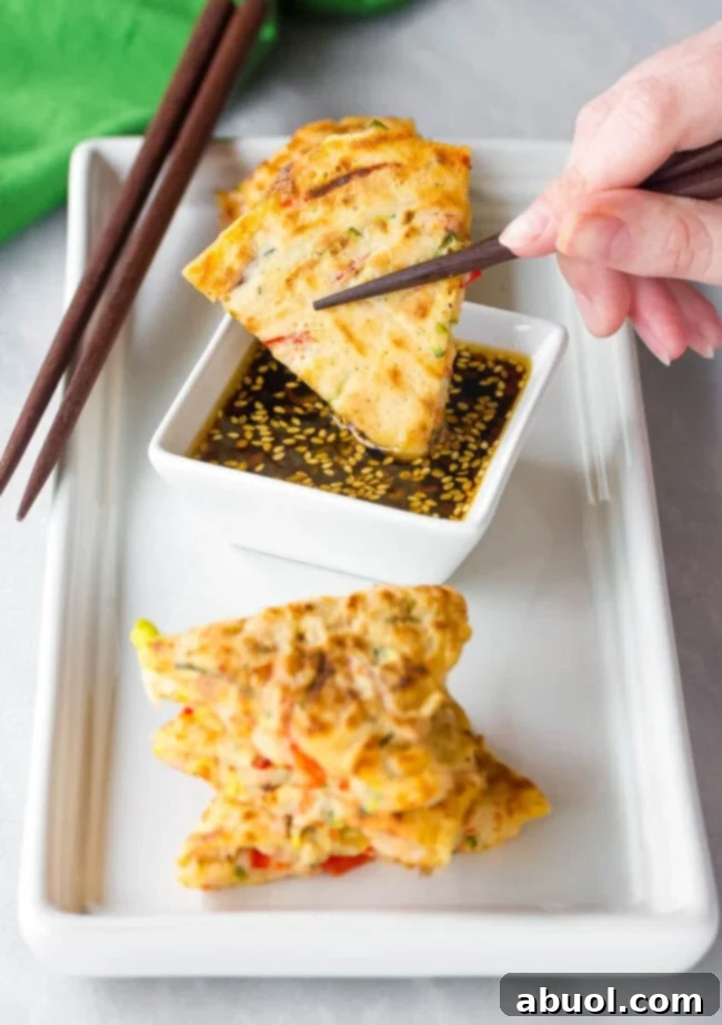 Korean Pancake Recipe #ASpicyPerspective #KoreanPancakes #KoreanPancakeRecipe #Pajun #Pajeon #Korean #Vegetarian A close-up shot of a hand expertly dipping a wedge of savory Korean vegetable pancake into a small bowl of spicy soy dipping sauce, highlighting the perfect pairing.
