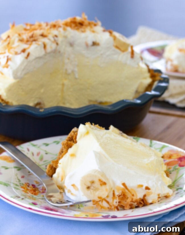A perfectly cut slice of Banana Cream Pie with a thick graham cracker crust, served on a plate with the whole pie in the background.