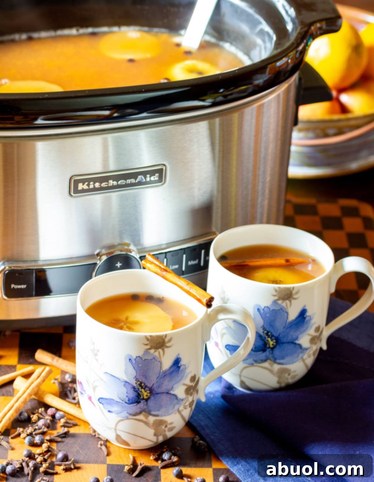 Two mugs brimming with hot apple cider wassail, set against a backdrop of a slow cooker filled with more spiced cider.