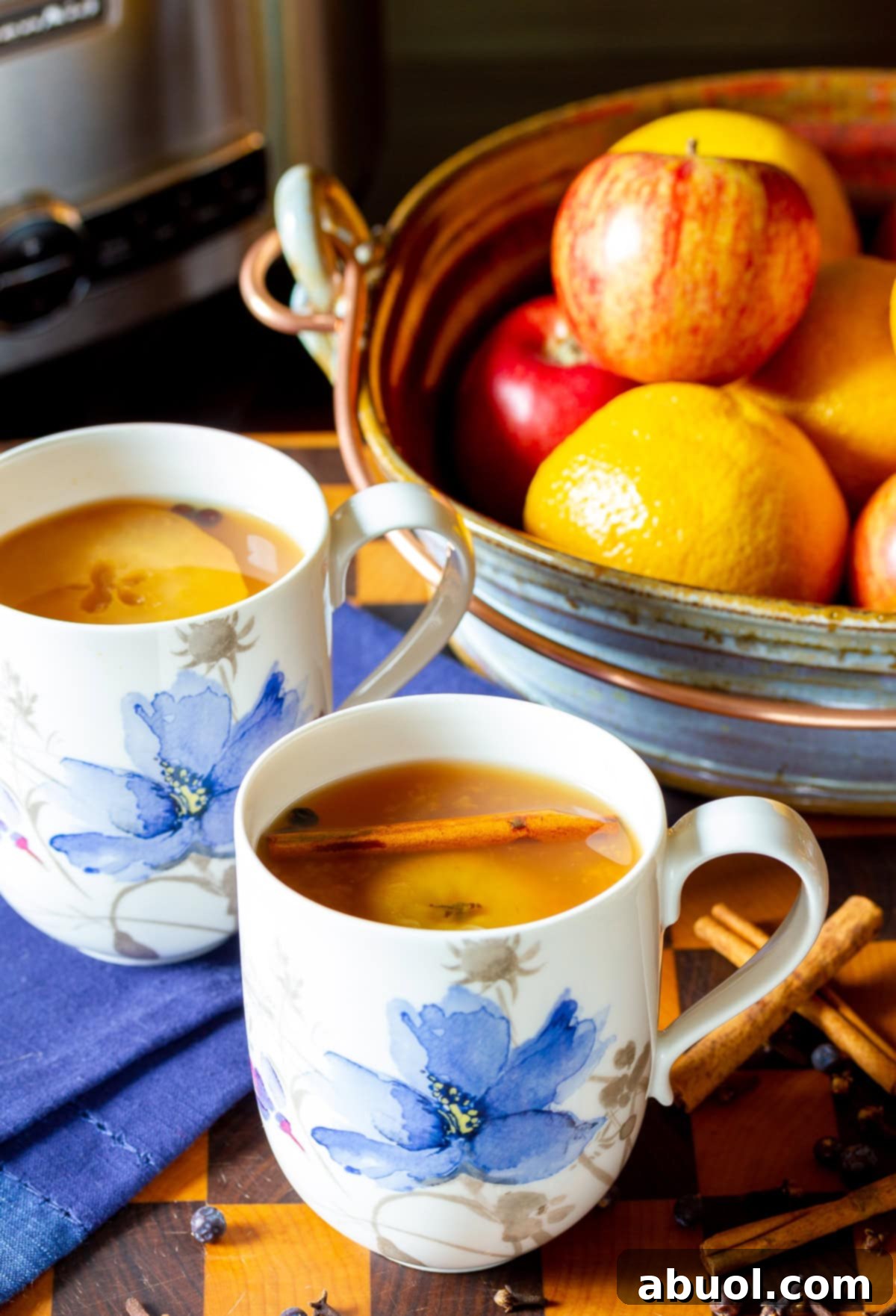 Two cozy mugs of apple cider wassail, resting beside a rustic bowl filled with fresh oranges and apples.
