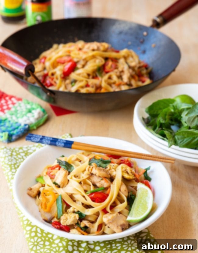 A steaming bowl of Drunken Noodles (Pad Kee Mao) served with fresh lime wedges and chopsticks, showcasing the rich colors of noodles, chicken, and vegetables, with another plate in the soft focus background.