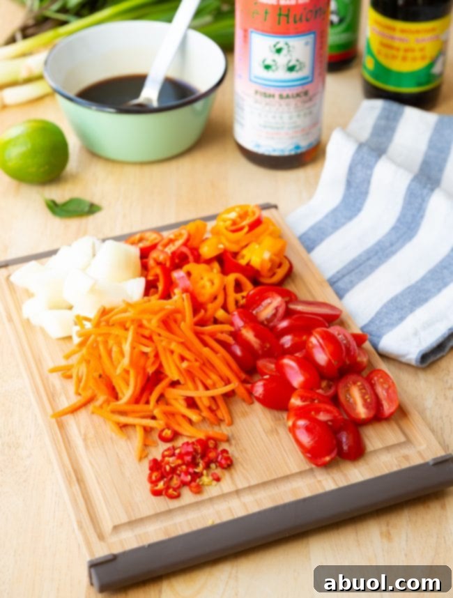 Assorted fresh vegetables for Pad Kee Mao, including sliced bell peppers, shredded carrots, chopped onions, whole Thai chilis, and fresh Thai basil leaves, neatly arranged on a wooden cutting board, ready for preparation.