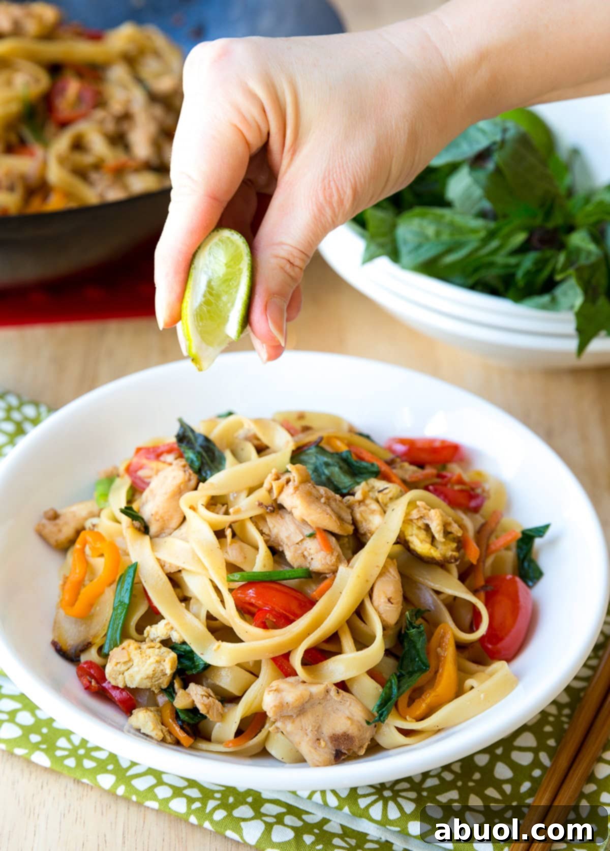 A hand is squeezing fresh lime juice over a generous serving of Pad Kee Mao in a bowl, enhancing the vibrant colors of the noodles and vegetables, with chopsticks on the side.