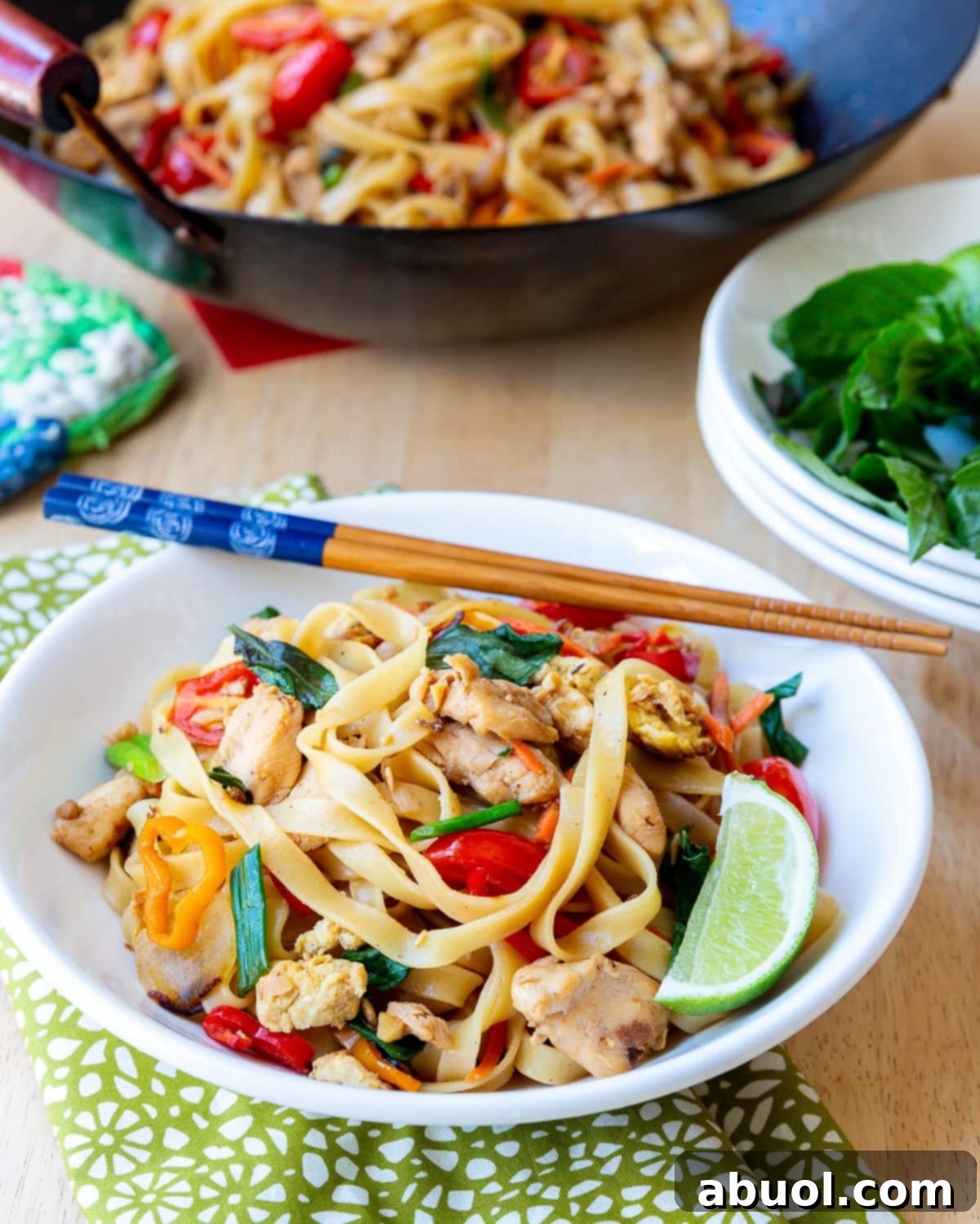 A vibrant bowl of Pad Kee Mao (Drunken Noodles) with chopsticks, garnished with a fresh lime wedge and a sprinkle of chopped green onions, presented against a rustic background.