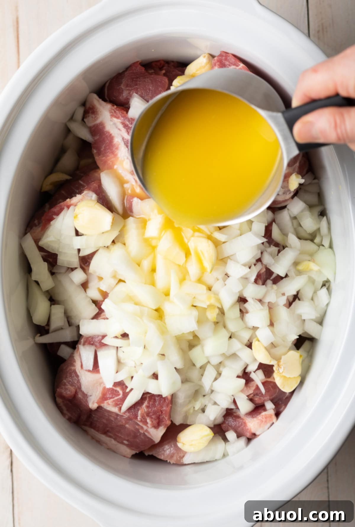 A hand pouring a cup of orange juice over pork, garlic, and onions in a slow cooker.