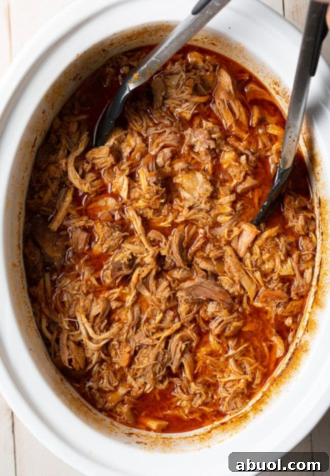 Tongs gently pulling apart tender shredded carnitas in the slow cooker.
