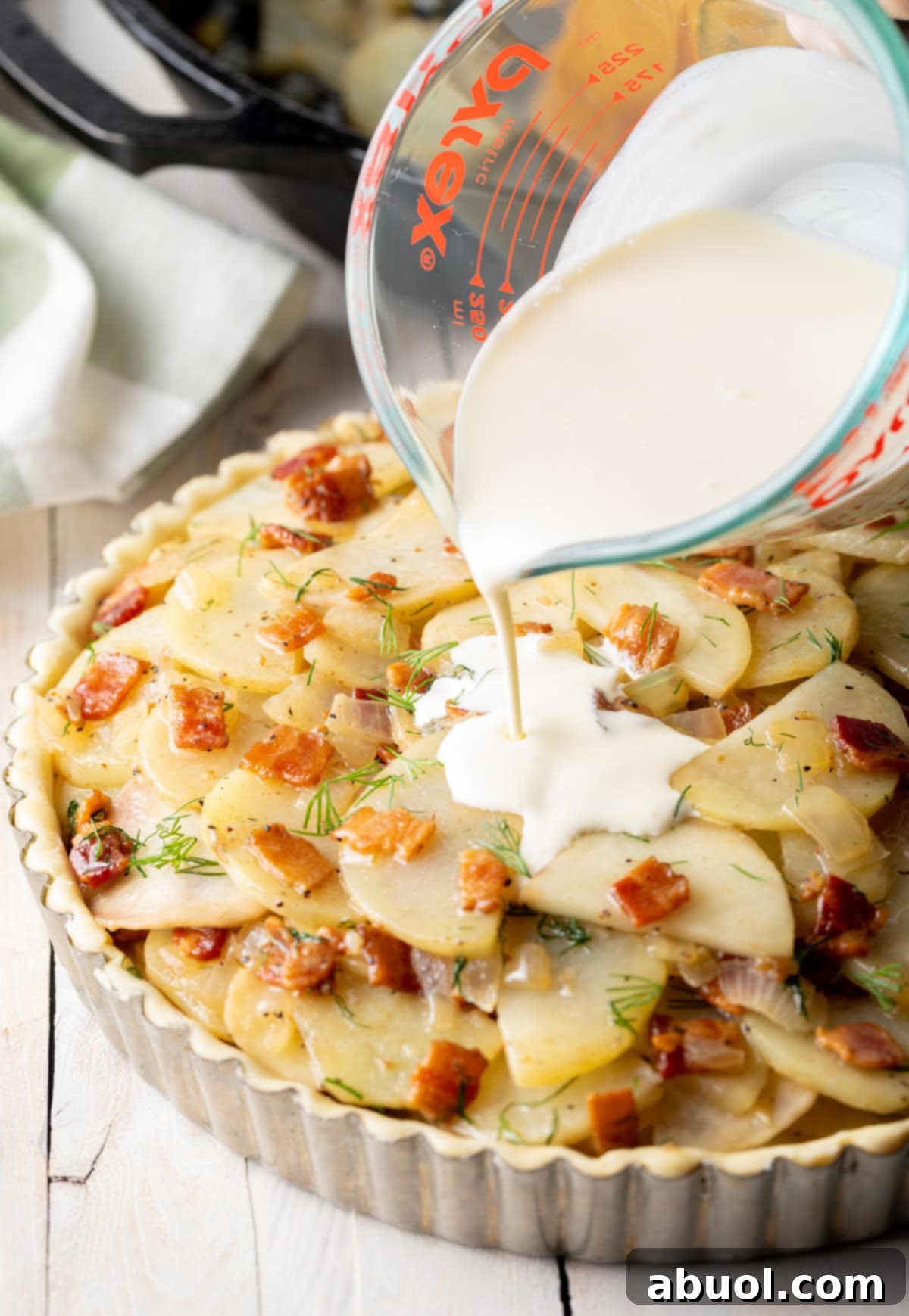 Heavy cream being poured generously over the top of layered potatoes and bacon in a tart tin, preparing for baking.