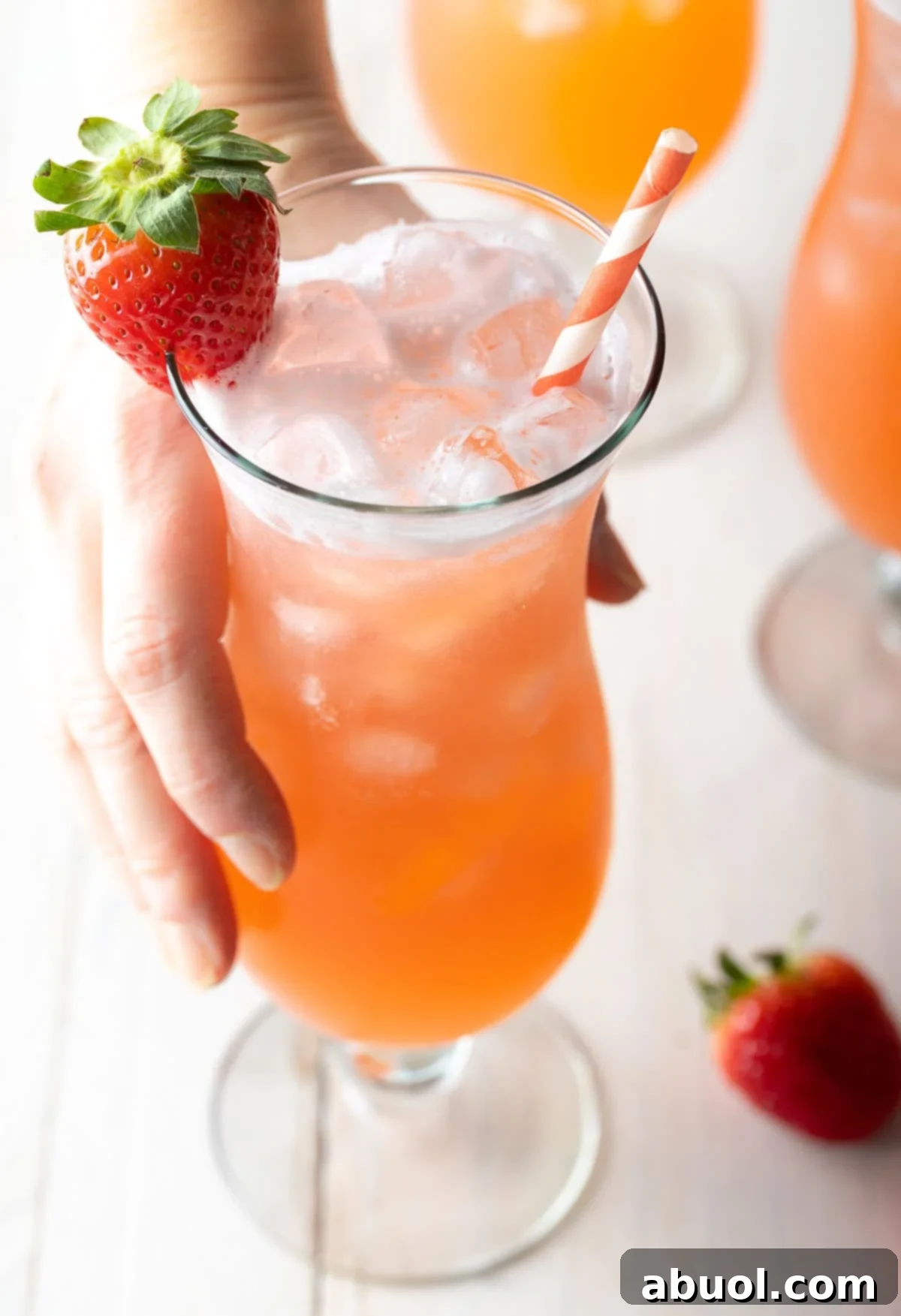Hand holding a tall cocktail glass filled with strawberry pineapple agua fresca. The glass is garnished with a strawberry on the rim and and orange and white striped paper straw.