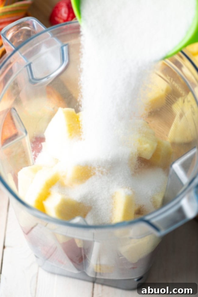 Pouring sugar into a blender pitcher with chunks of pineapple and strawberries.