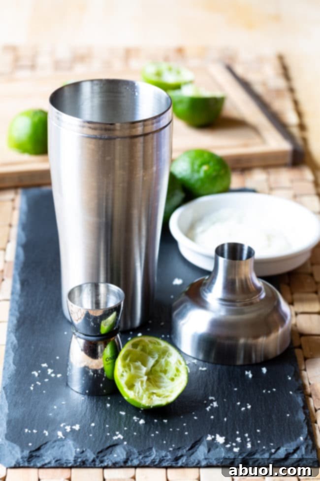 A cocktail shaker surrounded by fresh limes and a jigger, ready for margarita preparation.