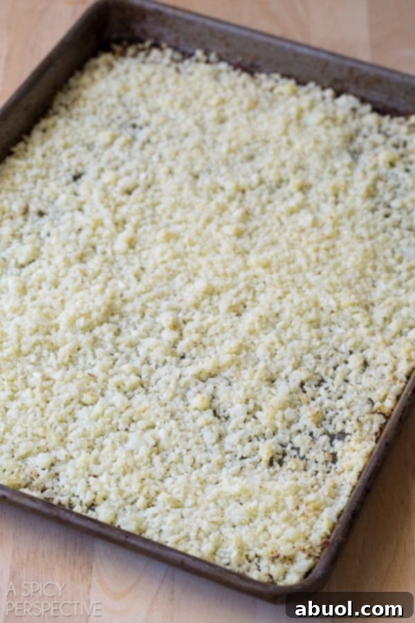 Riced cauliflower spread in a thin, even layer on a baking sheet, perfectly seasoned and ready for roasting.