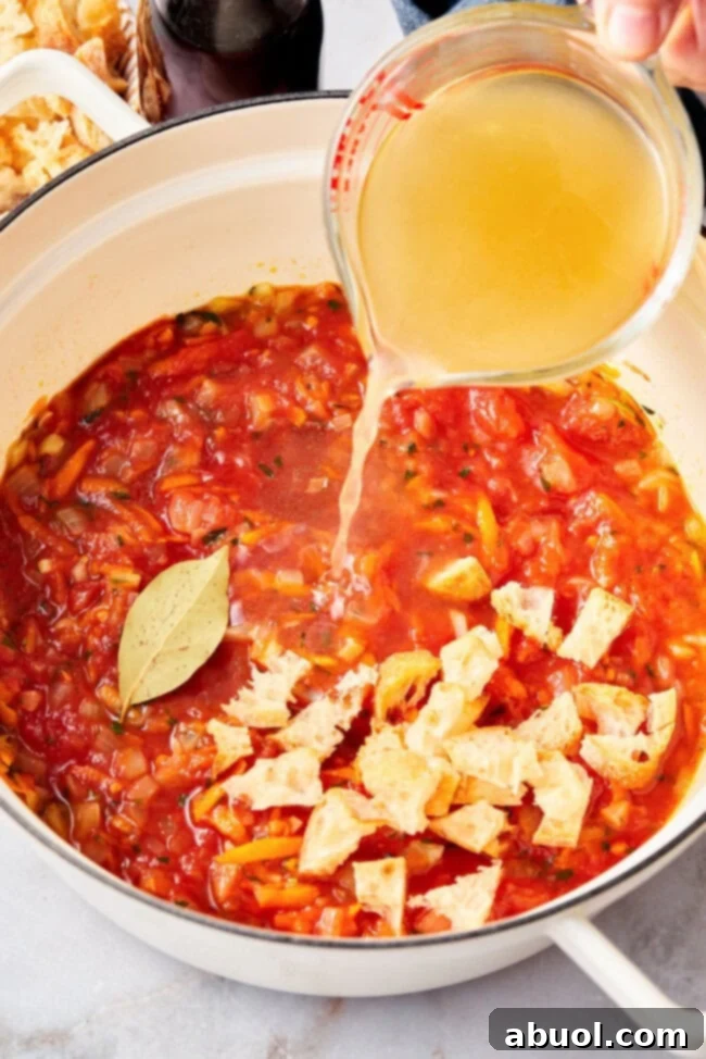 Broth being poured into a large stock pot with sautéed vegetables and tomatoes for Italian tomato soup.