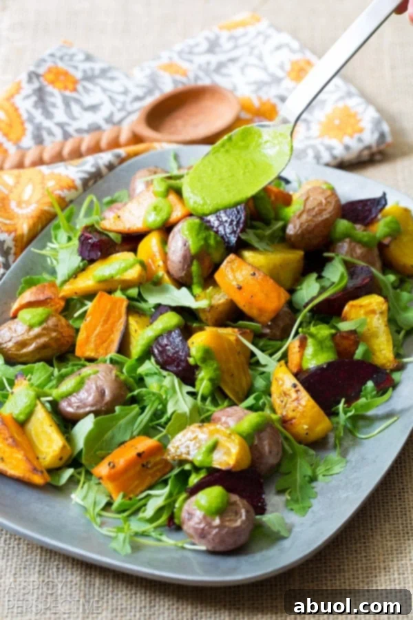 A hand drizzling homemade cilantro dressing over the roasted root salad on a serving platter, showing fresh green dressing over colorful vegetables.