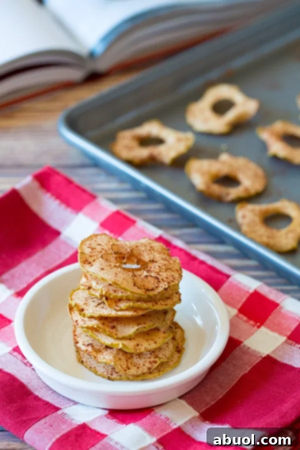 A stack of golden-brown baked apple chips in a white bowl, resting on a festive red and white linen, with more chips baking in the background.