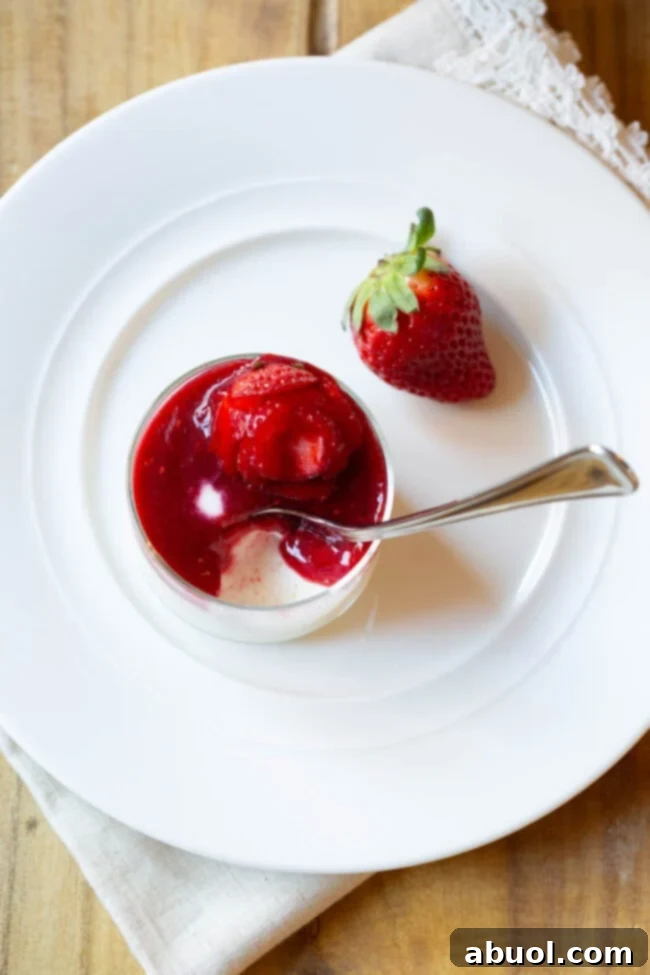 Top down view of a small glass dish with silky Swedish creme topped with berry compote and a strawberry rose, a spoon poised to scoop a bite.
