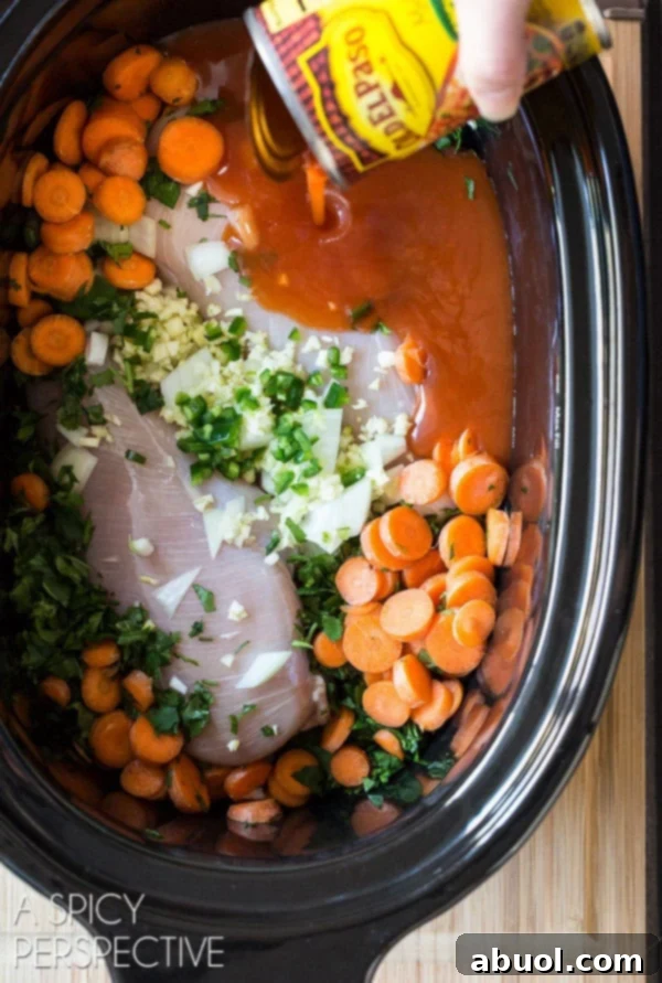 Top down view slow cooker filled with raw chicken breasts, fresh sliced carrots, onion, garlic, cilantro, and a hand adding enchilada sauce from a can.