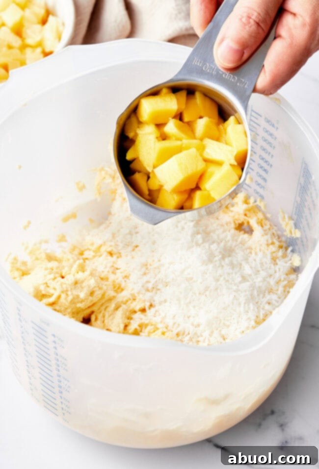 A hand holding a cup of fresh mango above the bowl of muffin batter.