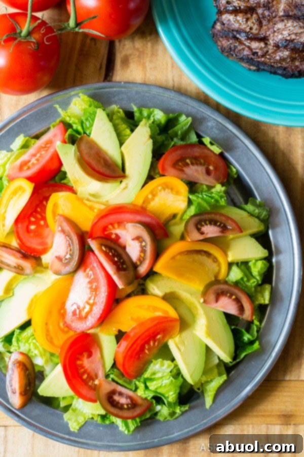 A prepared steak salad on a blue plate, garnished with fresh tomatoes and creamy avocado slices. Uncut steak and additional tomatoes are in the background, ready for preparation.