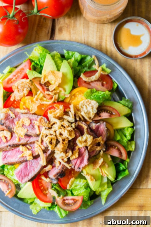 A blue plate holds a complete steak salad, perfectly arranged with vibrant tomatoes, creamy avocado, tender steak slices, and a generous sprinkle of crispy fried onions.