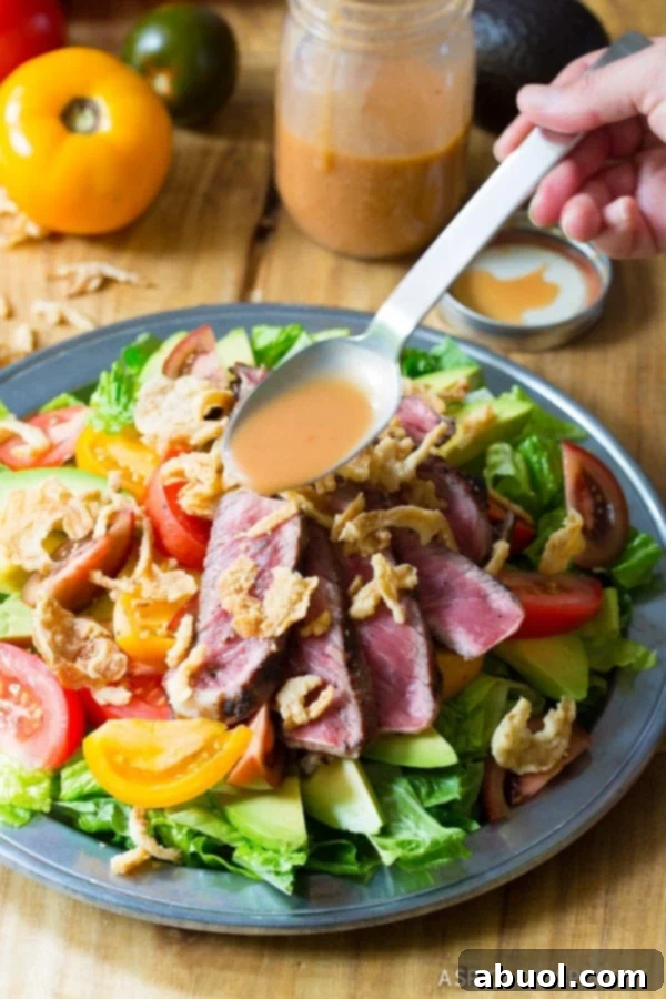 A hand holds a spoon, drizzling A1 vinaigrette over a beautifully plated steak salad, adorned with avocado, tomatoes, steak slices, and crispy fried onions.