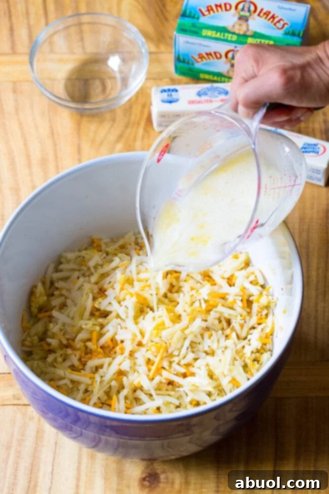 Melted butter being poured into a large mixing bowl containing shredded potatoes and shredded cheddar cheese.