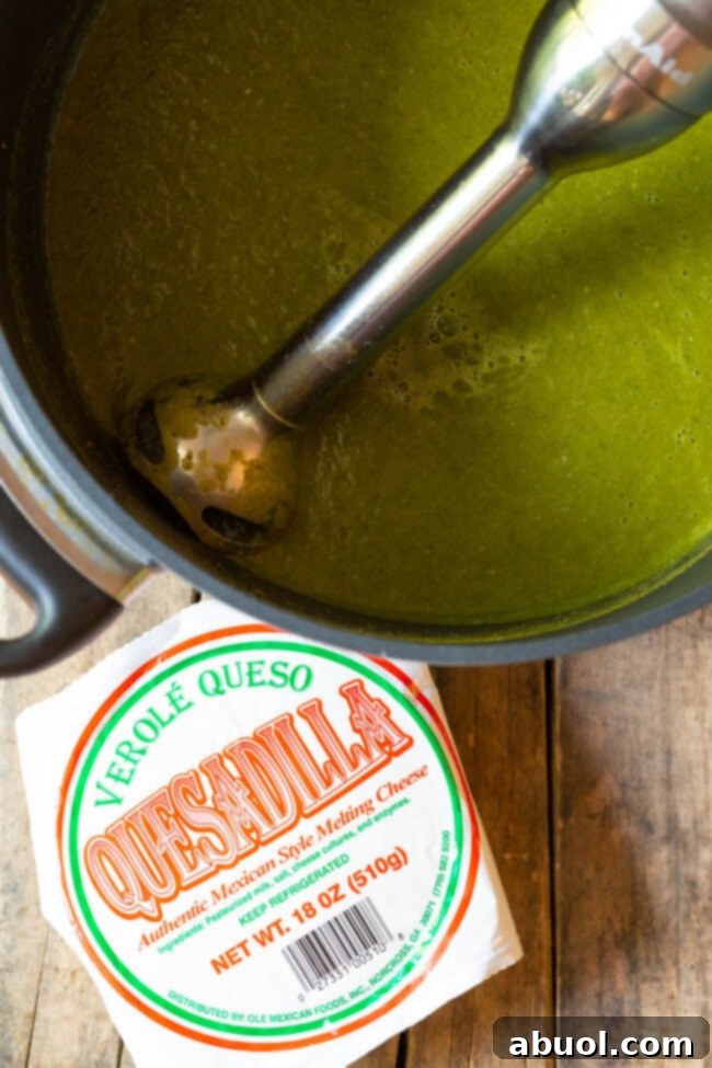 An immersion blender being used to create a creamy green soup in a large metal pot, with a block of Queso Quesadilla cheese resting beside the pot on a wooden surface.
