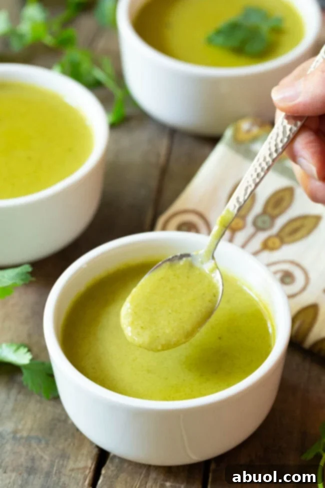A hand holding a metal spoon laden with creamy green soup, presented enticingly over a small white bowl, ready to be enjoyed.