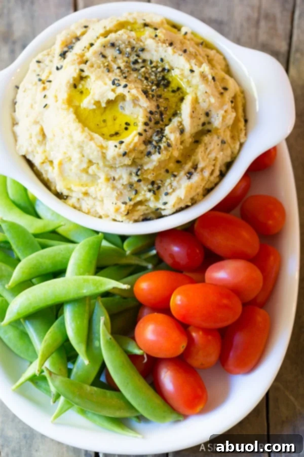 A vibrant plate featuring a white bowl of creamy sesame miso hummus, artfully garnished with sesame seeds, surrounded by fresh sweet peas and plump grape tomatoes. A delightful and healthy snack.