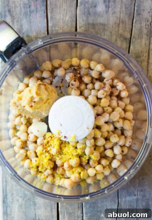 Top-down view of a food processor bowl filled with key ingredients for miso hummus: chickpeas, bright yellow lemon zest, a generous scoop of white miso paste, and whole cloves of garlic, ready for blending.