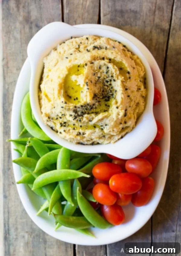 A beautifully presented plate of fresh sweet peas and vibrant grape tomatoes nestled around a gleaming white dish of creamy sesame miso hummus, ready for dipping and enjoying.