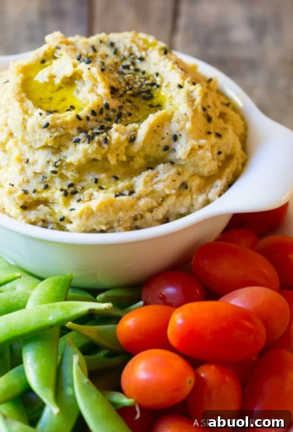 A close-up view of a pristine white bowl overflowing with beautifully smooth homemade miso paste hummus, elegantly garnished with a sprinkle of sesame seeds. Fresh sweet peas and vibrant grape tomatoes artfully arranged around the dish complete the inviting scene.