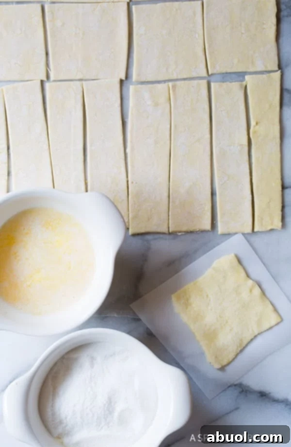 Raw puff pastry squares, melted butter, and granulated sugar in small bowls, demonstrating the ingredients for the Kouign Amann coating.