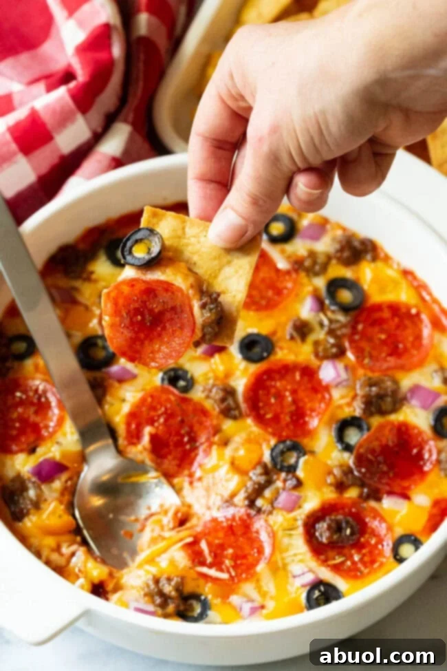 A hand expertly scoops a generous bite of the loaded Supreme Pizza Dip with a square pita cracker from a white round dish, showcasing the melted cheese, pepperoni, sausage, black olives, and red onion.