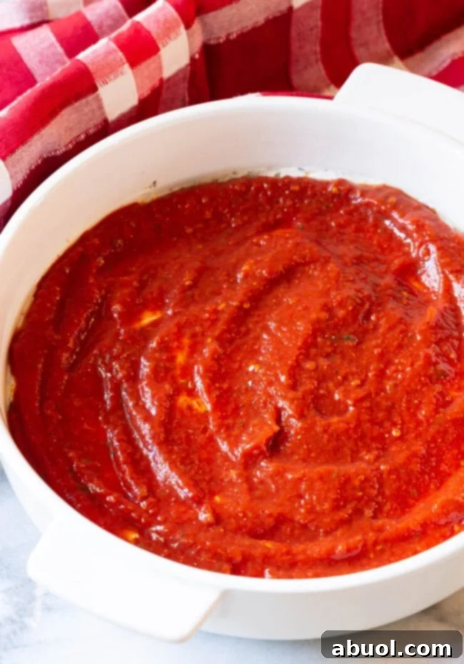 A close-up view of a white round baking dish, where a vibrant layer of tomato pizza sauce is perfectly spread over a creamy cheese base, ready for the next round of delicious toppings.